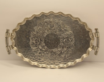 May include: An oval silver-toned metal tray with a scalloped edge and a hammered texture. The center features an embossed floral wreath design with roses and leaves. Two curved metal handles are attached to the sides with rivets.