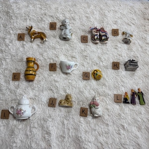 May include: A collection of miniature ceramic figurines and objects labeled A through L, including a dog, a child, clogs, a pitcher, a teacup, a hat, a box, a sugar bowl, a lion, a rabbit, and three small figures, all arranged on a textured surface.
