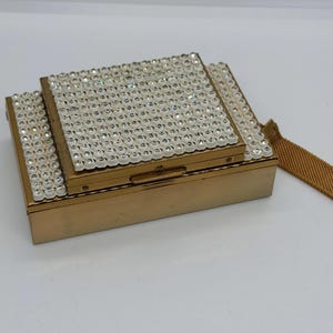 May include: A rectangular gold-toned metal clutch bag featuring a tiered design. The top surface is embellished with rows of clear, sparkling rhinestones. The clutch includes a matching gold-toned metal mesh wrist strap attached to the side.