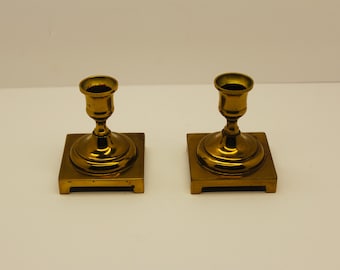 May include: A pair of polished brass candle holders, each featuring a cylindrical socket atop a rounded pedestal base, which is mounted on a square, flat platform with four small feet at the corners, creating a symmetrical metallic set.