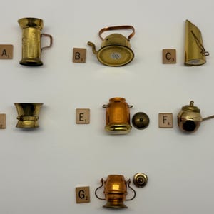 May include: A collection of miniature brass kitchenware, including a pitcher, kettle, and pot. The items are arranged on a white surface, with Scrabble tiles next to each piece. The brass items have a vintage look.