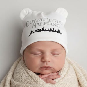 Op de afbeelding: Een pasgeboren baby met een witte muts met berenoren en de grijze tekst "Cutest Little Halfling". De baby is gewikkeld in een crèmekleurige gebreide deken.