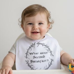 Op de afbeelding: Witte baby slabbetje met de zwarte tekst "What about Second Breakfast?", omringd door een cirkelvormig ontwerp. Het slabbetje wordt gedragen door een lachende baby in een kinderstoel.