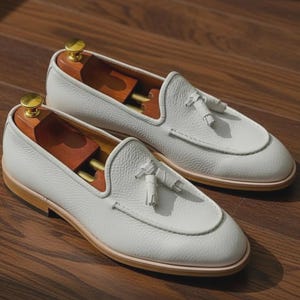 May include: A pair of white leather loafers with a textured finish. Each shoe features a decorative tassel and a light brown sole. Wooden shoe trees are inserted inside the shoes. The shoes are displayed on a wooden surface.