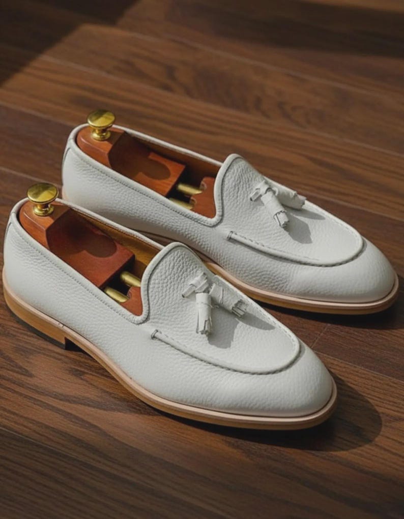 May include: A pair of white leather loafers with a textured finish. Each shoe features a decorative tassel and a light brown sole. Wooden shoe trees are inserted inside the shoes. The shoes are displayed on a wooden surface.