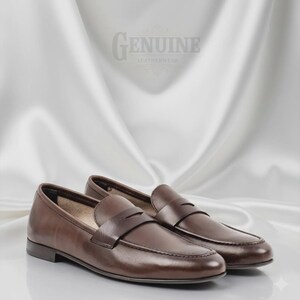 Op de afbeelding: Een paar bruine leren penny loafers. De schoenen hebben een klassiek ontwerp met een ronde neus, een band over de wreef en een lage hak. De achtergrond is een witte satijnen stof met de tekst "GENUINE LEATHERWEAR".