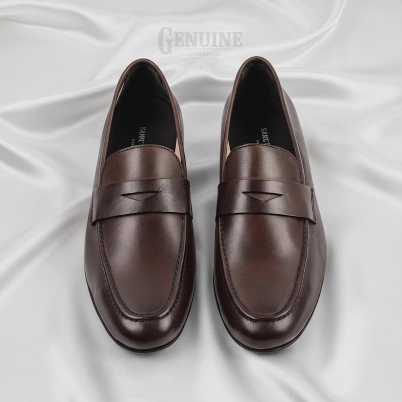 Op de afbeelding: Een paar bruine leren loafers met een klassiek ontwerp. De schoenen hebben een gladde, gepolijste afwerking en een banddetail over de bovenkant. Het woord "GENUINE" staat bovenaan de afbeelding.