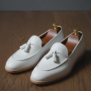 May include: A pair of white leather loafers with tassel details. The shoes have a rounded toe and a light brown sole. The loafers are displayed on a wooden surface.