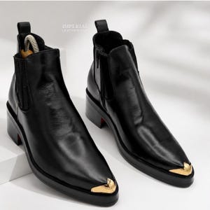 Puede incluir: Un par de botines Chelsea de cuero negro con punta afilada y una puntera de metal dorado pulido. Los botines tienen paneles laterales elásticos, una lengüeta en el talón y un tacón bajo en bloque. El texto IMPERIAL LEATHER HUB es visible.