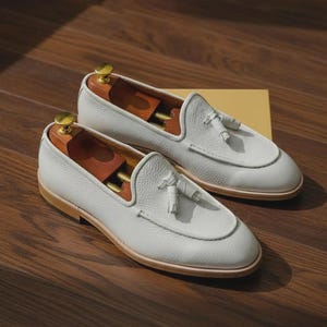 May include: A pair of white leather loafers with tassel details. The shoes have a rounded toe and a light brown sole. Wooden shoe trees are inside the shoes. The loafers are displayed on a wooden surface.