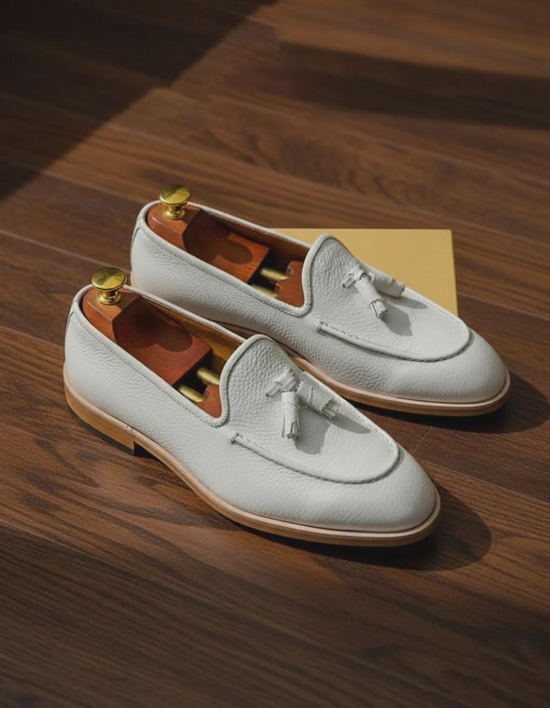 May include: A pair of white leather loafers with tassel details. The shoes have a rounded toe and a light brown sole. Wooden shoe trees are inside the shoes. The loafers are displayed on a wooden surface.