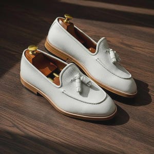 May include: A pair of white leather loafers with tassel details. The shoes have a rounded toe and a light brown sole. Shoe trees are inserted inside the shoes. The loafers are displayed on a dark wood surface.
