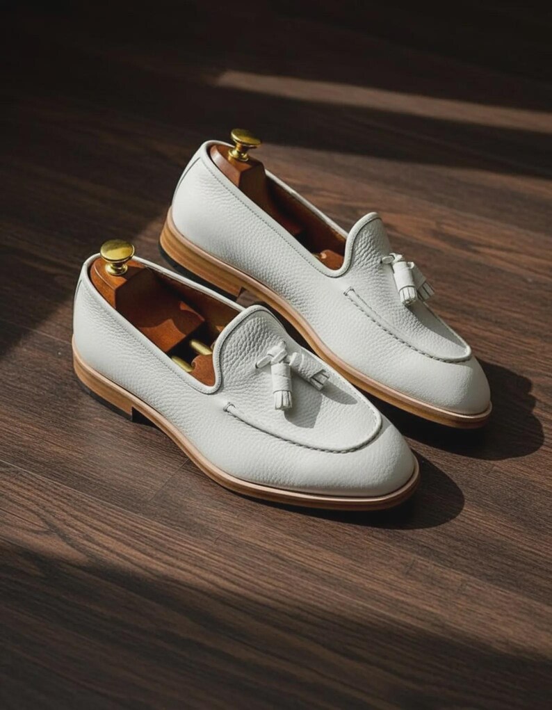 May include: A pair of white leather loafers with tassel details. The shoes have a rounded toe and a light brown sole. Shoe trees are inserted inside the shoes. The loafers are displayed on a dark wood surface.
