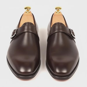 Könnte beinhalten: Ein Paar braune Leder-Monk-Schuhe mit silbernen Schnallen. Die Schuhe haben eine polierte Oberfläche und werden mit hölzernen Schuhspannern präsentiert. Klassisches Design.