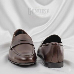 Op de afbeelding: Een paar bruine leren loafers met een klassiek design. De schoenen hebben een gladde, gepolijste afwerking en een band over de bovenkant. Het woord "GENUINE LEATHERWEAR" is zichtbaar op de achtergrond.