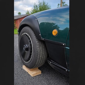 May include: A close-up of a dark green car's front wheel and fender. The wheel is black with a silver rim and is resting on a wooden block. The car's body has black trim and a yellow side marker light.