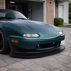 Puede incluir: Un coche deportivo verde oscuro con detalles en negro. Se muestra la parte delantera del coche, con faros, un labio delantero negro y un área para la matrícula. El coche está estacionado en un camino pavimentado con ladrillos.