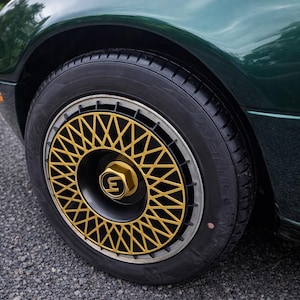 Op de afbeelding: Close-up van een autowiel met een zwarte band en een goudkleurige en zwarte velg. De velg heeft een geometrisch patroon en een gouden naafkap met een gestileerd "E"-logo. De carrosserie van de auto is donkergroen.