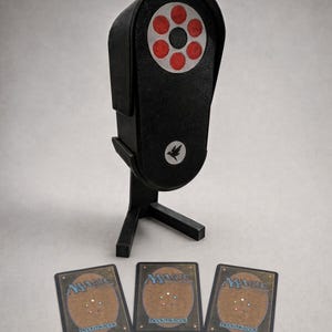 Pode incluir: Um suporte para cartas preto de formato oval com uma seção superior circular apresentando seis detalhes circulares vermelhos e um pequeno emblema de pássaro. Três cartas colecionáveis Magic: The Gathering estão dispostas em fila à frente do suporte, mostrando o verso.