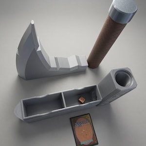 May include: A grey 3D-printed axe-shaped storage case with a brown textured handle, featuring a hollow interior divided into two compartments. A single small wooden die sits inside one section, and a Magic: The Gathering trading card lies nearby.