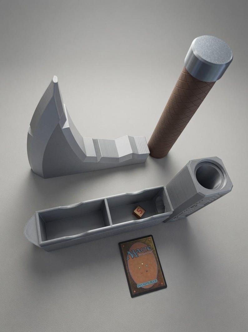 May include: A grey 3D-printed axe-shaped storage case with a brown textured handle, featuring a hollow interior divided into two compartments. A single small wooden die sits inside one section, and a Magic: The Gathering trading card lies nearby.