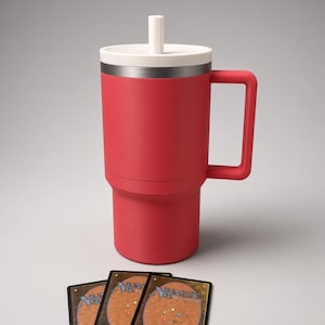 Puede incluir: Un vaso térmico rojo con asa cuadrada, tapa blanca y pajita blanca. El vaso tiene un borde plateado metálico. Tres cartas coleccionables de Magic: The Gathering con el texto Deckmaster están colocadas delante del vaso.