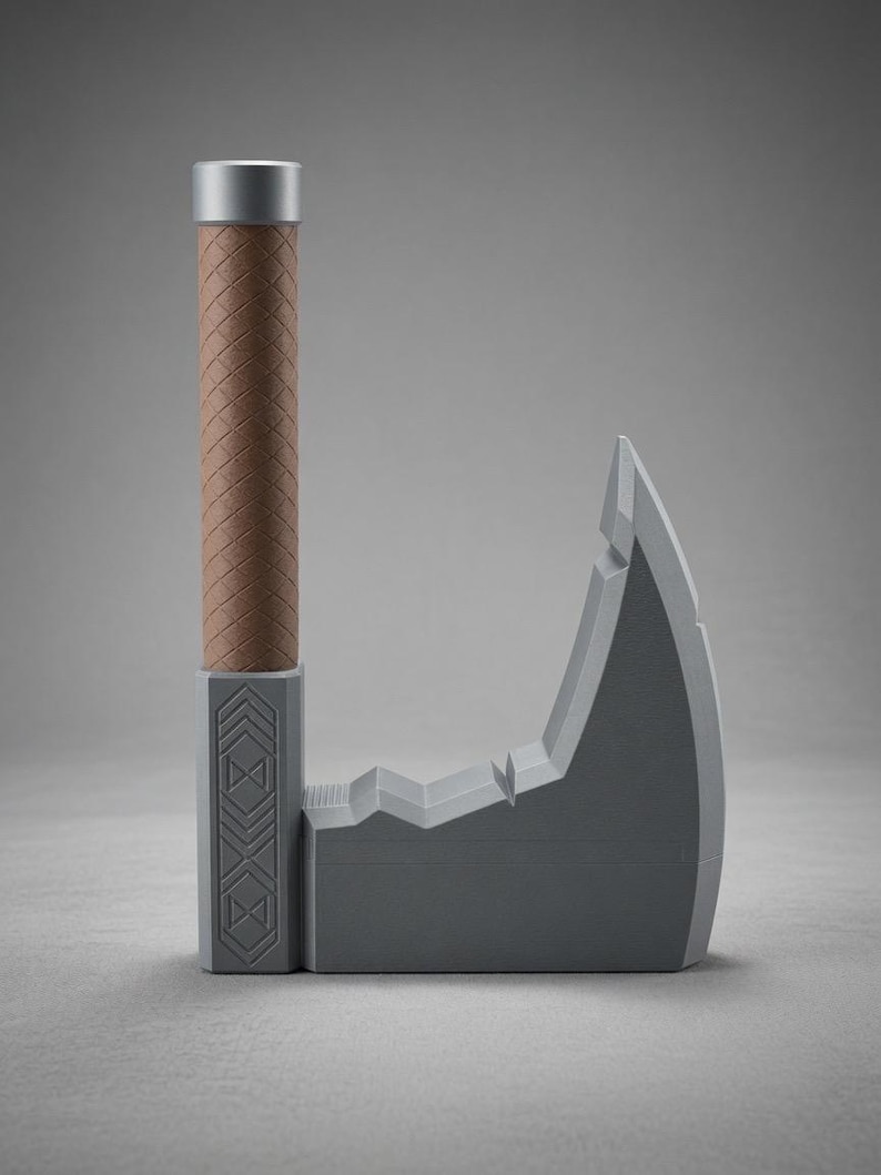 May include: A grey axe with a textured brown cylindrical handle and a silver cap. The axe head features a geometric pattern of stacked triangles and a stepped, notched top edge. The design is symmetrical and stands upright on a flat base.