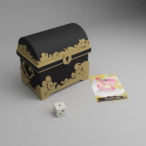 Könnte beinhalten: Eine schwarze Schatzkiste mit goldenen Verzierungen, ein weißer Würfel und eine Sammelkarte. Die Truhe hat ein Schlüsselloch und dekorative wolkenartige Designs. Die Karte zeigt eine Figur mit rosa Haaren.