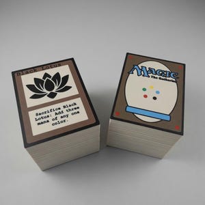 May include: Two stacks of Magic: The Gathering trading cards. One stack shows the "Black Lotus" card with a black lotus flower graphic and text. The other stack shows the card back with the word "Magic" and a white oval.
