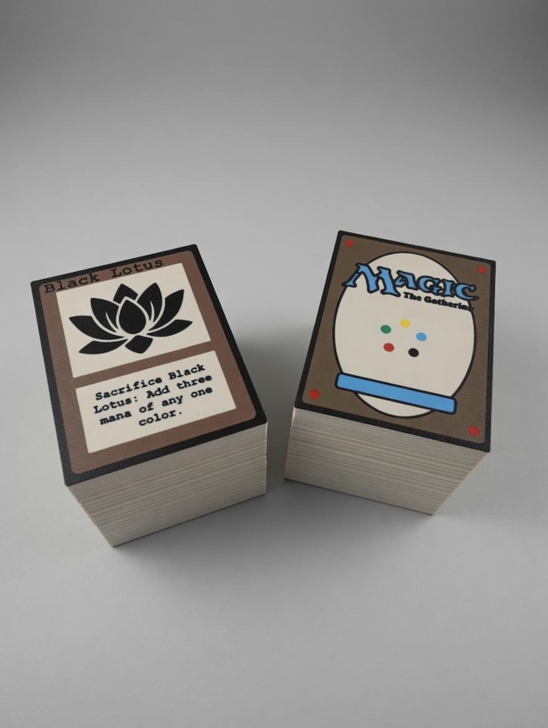 May include: Two stacks of Magic: The Gathering trading cards. One stack shows the "Black Lotus" card with a black lotus flower graphic and text. The other stack shows the card back with the word "Magic" and a white oval.