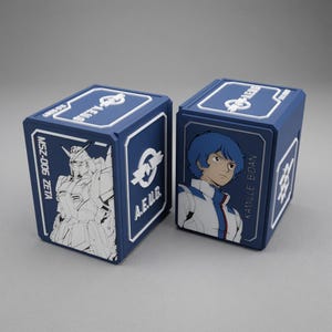 Puede incluir: Dos cajas de almacenamiento rectangulares azules con detalles en relieve blancos. Una caja muestra una ilustración lineal del robot MSZ-006 Zeta y el texto A.E.U.G. La otra caja presenta un retrato de Kamille Bidan y el logotipo A.E.U.G.