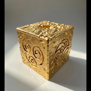 May include: A gold-colored, cube-shaped decorative box with a textured surface. The box features carved spiral and wave-like designs on its sides. A slot is visible on the top surface. The box is likely made of metal or a similar material.