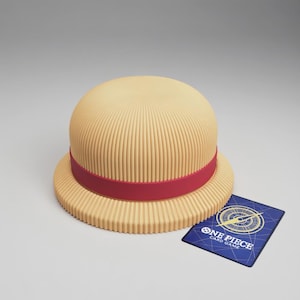 Op de afbeelding: Een bruin strooien hoed met een rode band, naast een blauwe One Piece kaart. De hoed heeft een afgeronde kroon en een brede rand met verticale ribbels. De kaart heeft een kompasontwerp.