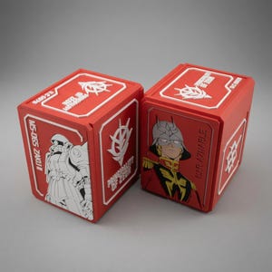 Könnte beinhalten: Zwei rote, rechteckige Aufbewahrungsboxen mit weißen Prägedetails. Eine Box zeigt einen Zaku II Roboter und den Text MS-065 ZAKU II, die andere ein Charakterporträt mit der Aufschrift CHAR AZNABLE. Beide tragen das Emblem von Zeon.