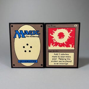 Peut inclure: Deux cartes à collectionner Magic: The Gathering. L'une présente le logo du jeu et le mot "DECKMASTER". L'autre est intitulée "Sol Ring" avec des graphismes rouges et jaunes et du texte décrivant sa fonction.