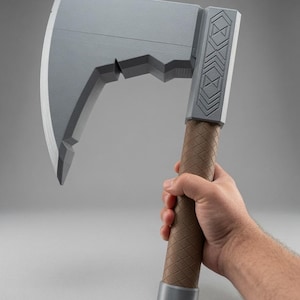 Puede incluir: Un hacha gris con una hoja ancha y curva de borde dentado. El mango es marrón con una textura de rejilla, y la cabeza presenta una sección rectangular con patrones geométricos grabados. La base del mango tiene una tapa gris lisa.