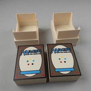 May include: Two stacks of Magic: The Gathering playing cards with the card backs visible, featuring the game's logo and a brown border. Two beige card storage boxes are positioned above the card stacks, ready for use.