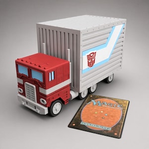 May include: A red and white toy truck with a large rectangular trailer featuring a blue stripe and a red Autobot symbol. Beside the truck lies a rectangular Magic The Gathering Deckmaster trading card with an orange and brown back design.
