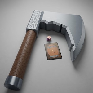 May include: A silver-coloured toy axe with a brown, diamond-patterned handle and geometric engravings on the head. Beside the axe lies a small pink six-sided die showing the number one and a rectangular Magic: The Gathering trading card.