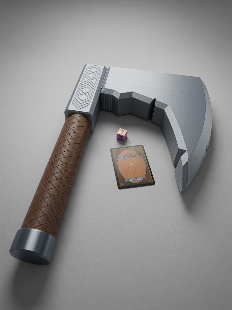 May include: A silver-coloured toy axe with a brown, diamond-patterned handle and geometric engravings on the head. Beside the axe lies a small pink six-sided die showing the number one and a rectangular Magic: The Gathering trading card.