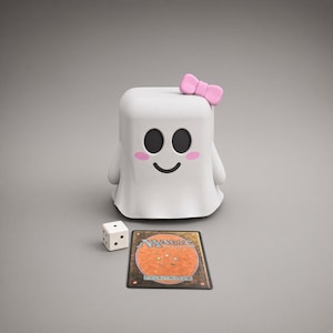 May include: A white ghost-shaped deck box featuring a pink bow on top, black oval eyes, a curved black mouth, and pink circular cheeks. Beside the box sits a white six-sided die with black pips and a Magic: The Gathering trading card.