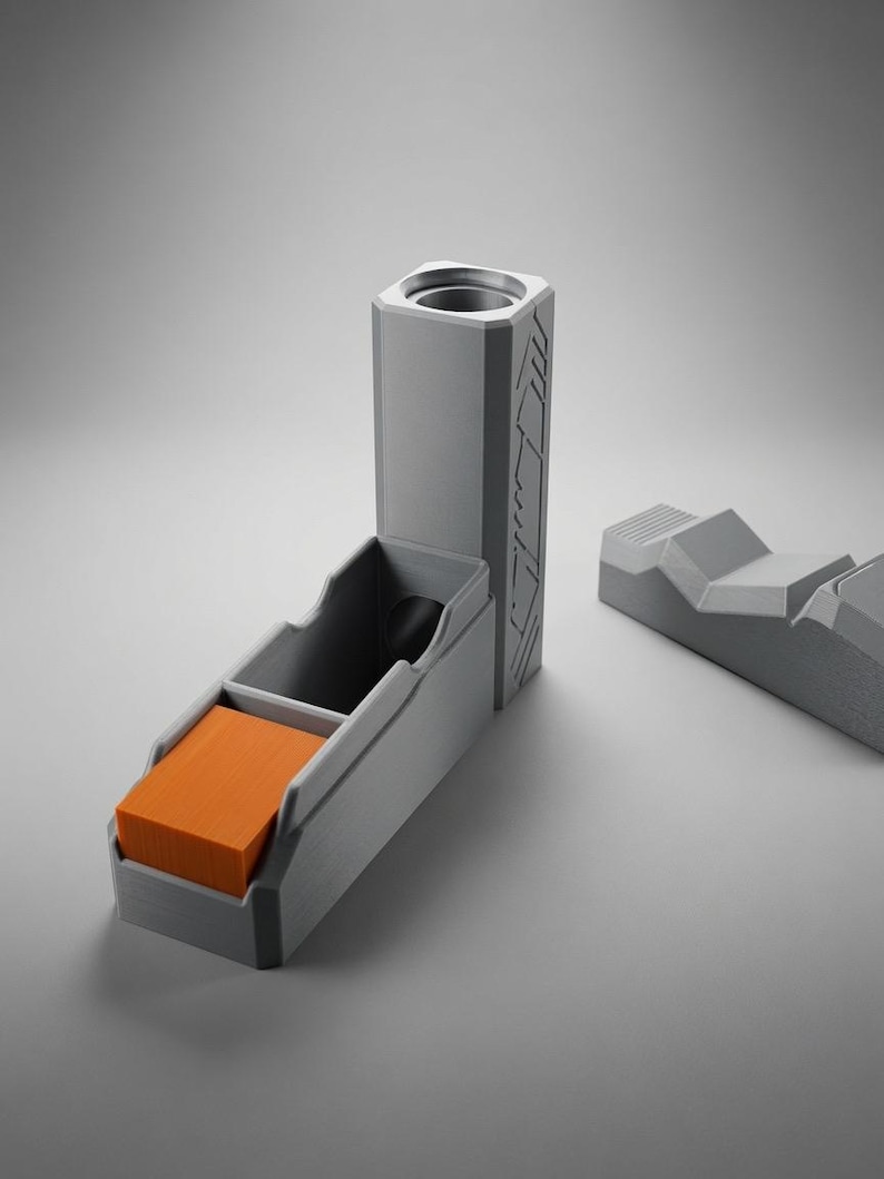 May include: A grey 3D-printed dice tower with a vertical cylindrical opening and a rectangular tray containing a stack of orange square tiles. A separate grey geometric piece with angled ridges sits to the right of the main tower structure.