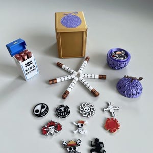 May include: A selection of anime-themed accessories, including a gold box with a purple fruit design, a pack of cigarettes with the text "KING GROUND", and various skull and crossbones pins. The items are arranged on a light grey surface.