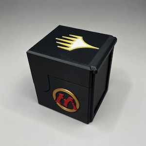 May include: Black, cube-shaped card storage box with gold and red accents. The box features a gold emblem on the lid and a red and gold emblem on the front, likely for collectible card games.
