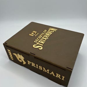 Può includere: Una scatola marrone con scritte dorate e un logo. Il testo sulla scatola recita "Secrets of Strixhaven" e "Prismari". La scatola è probabilmente per riporre carte o altri oggetti da collezione.