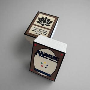 May include: Two stacks of Magic: The Gathering playing cards. The top card of one stack features a black lotus flower graphic and text. The other stack shows the back of the cards with the game's logo and colorful dots.