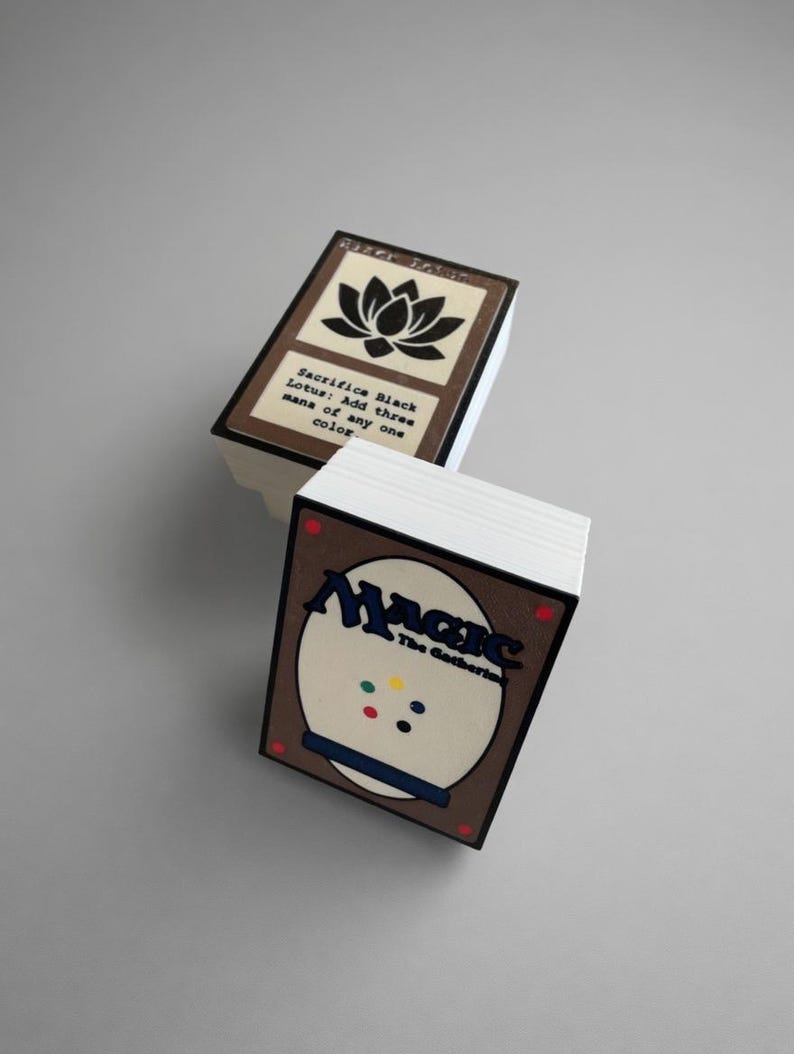 May include: Two stacks of Magic: The Gathering playing cards. The top card of one stack features a black lotus flower graphic and text. The other stack shows the back of the cards with the game's logo and colorful dots.