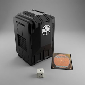 May include: A black, rectangular, textured deck box featuring a white circular tree emblem on the front. Beside the box lies a white six-sided die with black pips and a single Magic the Gathering trading card with a tan back and coloured dots.