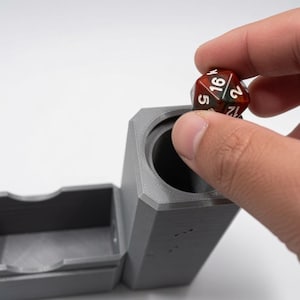 May include: A matte grey, 3D-printed cylindrical dice tower with an attached rectangular tray. A multi-coloured, translucent red and brown twenty-sided die with white numbering is being placed into the top opening of the tower.