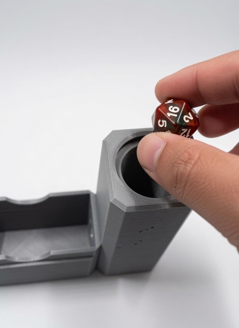 May include: A matte grey, 3D-printed cylindrical dice tower with an attached rectangular tray. A multi-coloured, translucent red and brown twenty-sided die with white numbering is being placed into the top opening of the tower.
